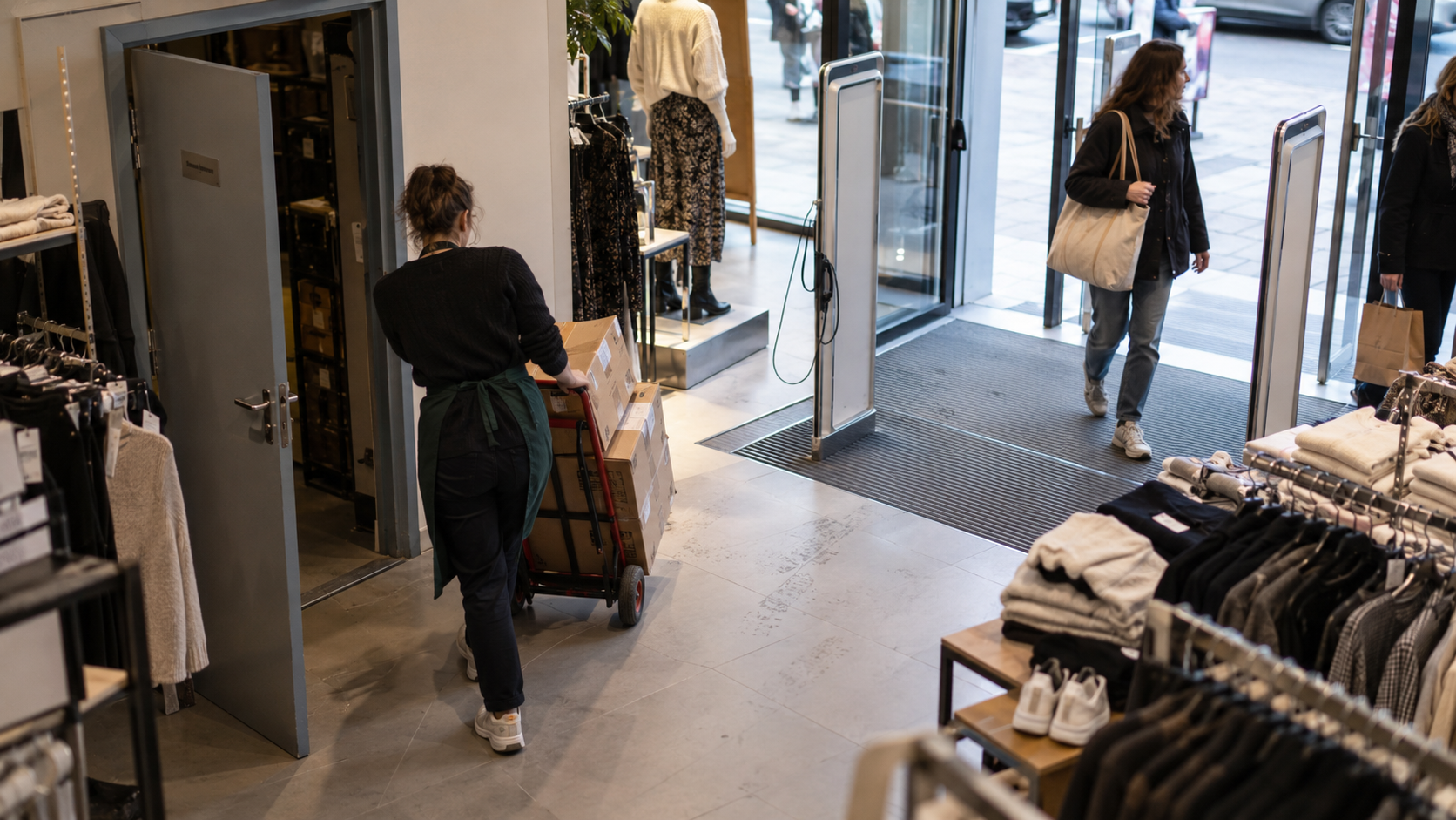 Vue en plongée de l’entrée d’un magasin de vêtements : une vendeuse déplace des cartons sur un diable près de la réserve, tandis que deux clientes entrent par les portes vitrées.