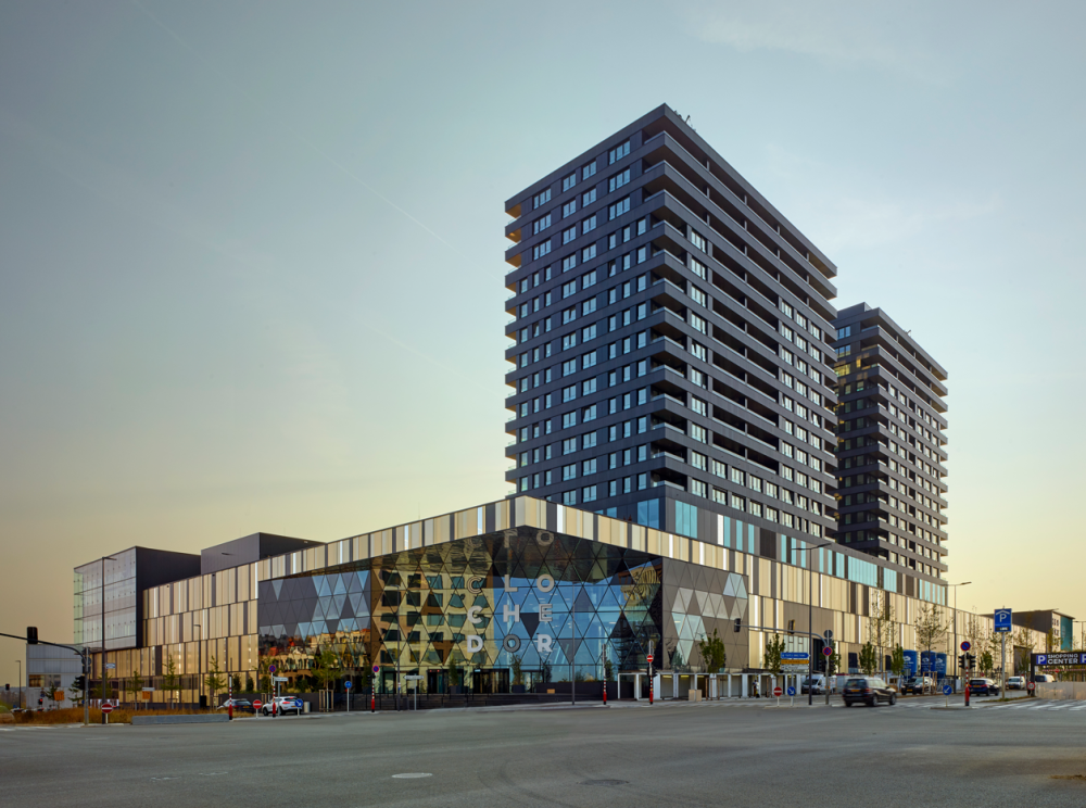 Vue extérieure du centre commercial La Cloche d'Or, un bâtiment moderne à l’architecture contemporaine, surmonté de deux tours résidentielles. Le ciel est dégagé et l’esplanade devant le bâtiment est vide.