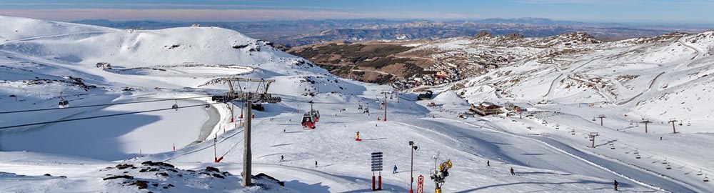 Vue panoramique de la station de ski de Sierra Nevada sous un ciel bleu, avec des skieurs sur les pistes enneigées et les remontées mécaniques en arrière-plan.